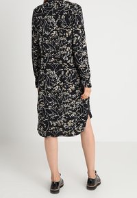 Musta kukkamekko pitkähihaisilla, jossa on valkoinen ja beige kukkakuosi. Kangas on pehmeä ja hieman kiiltävä, ja siinä on sivuventtiilit. Mustat kengät.