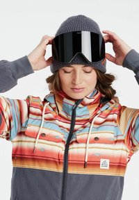 Pestrý snowboardový kabát so štrkovanými pruhmi v červených, oranžových, modrých a šedých odtieňoch. Obsahuje zips, vrecká a pletenú šedú čiapku s okuliarmi.