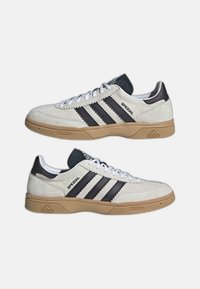 Baskets en suède blanc avec accents en cuir noir, trois bandes noires, et semelles en caoutchouc naturel. Présentent un design à lacets et des détails texturés.