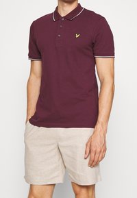 Burgundy pikétröja med krage, tre-knapps placket och broderad logotyp. Har grå trim på ärmar och krage. Bärs med beigea shorts.