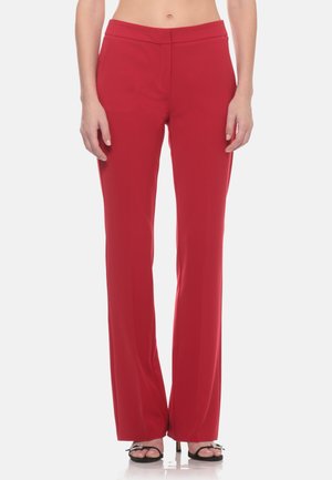 Donna con pantaloni rossi a vita alta e gamba dritta e tacchi neri open toe con cinturini alla caviglia, in piedi su uno sfondo semplice.