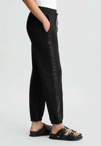 Pantalons de survêtement noirs avec taille élastique, poches latérales et rayure verticale texturée contrastante. Associés à des sandales glissantes noires.