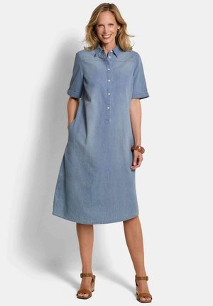 Frau lächelt und trägt ein hellblaues Denim-Midikleid mit kurzen Ärmeln, Taschen, braunem Armband und braunen Sandalen mit Absatz.