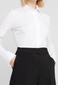 Žena ve fitted bílé košili s dlouhým rukávem, zastrčené do černých kalhot s vysokým pasem, s rukama v kapsách.