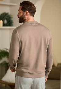 Sweatshirt marron clair en tissu doux, avec un col rond et des poignets et un ourlet côtelés. Porté avec un pantalon de jogging gris dans un cadre neutre.