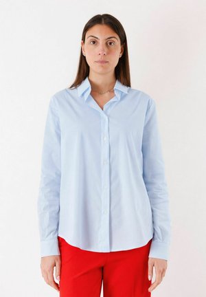 Donna con capelli lunghi e scuri che indossa una camicia azzurro chiaro abbottonata e pantaloni rossi, in piedi davanti a uno sfondo bianco uniforme.