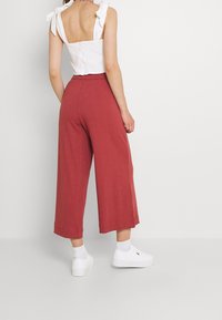 Femme portant un crop top sans manches blanc avec des épaules nouées, un pantalon large taille haute de couleur rouille, des chaussettes blanches et des baskets blanches, se tenant de profil.
