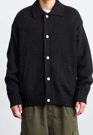 Mand iført sort strikket cardigan med knapper, ribbede manchetter og krave, kombineret med olivengrønne cargobukser, stående foran en ensfarvet baggrund.