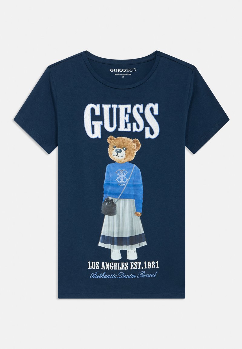 Μπλε ναυτικό μπλουζάκι με γραφικό αρκούδας σε μπλε πουλόβερ και φούστα, με το κείμενο "GUESS" πάνω και "LOS ANGELES EST. 1981" κάτω.