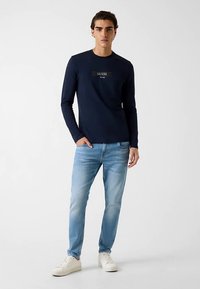 Chemise à manches longues bleu marine avec un logo central, associée à un jean bleu clair et des baskets blanches. La tenue présente un design décontracté et ajusté.