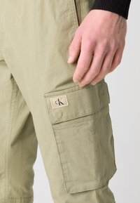 Cargo broek in lichtgroene stof met een gestructureerde afwerking. Voorzien van een zijzak met een beige label waarop "ck" staat.