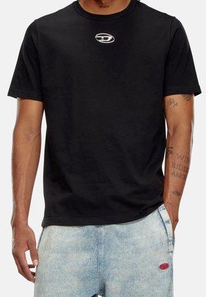 Homme portant un t-shirt noir à manches courtes avec un petit logo ovale sur la poitrine et un jean bleu clair délavé avec un patch ovale rouge sur la poche.