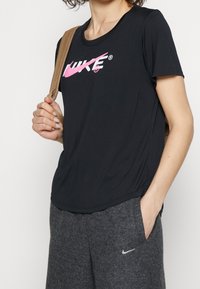 Camiseta negra de manga corta de Nike con un logo rosa prominente. Combinada con pantalones deportivos grises, sostenidos con una correa beige en el hombro.