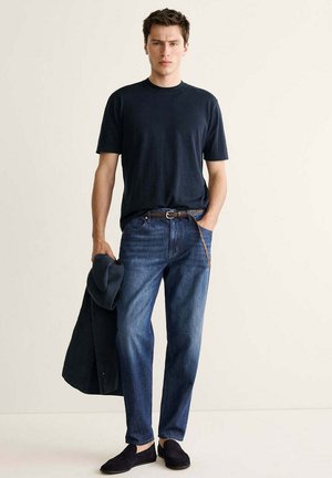 Un homme debout porte un t-shirt bleu marine, un jean bleu, une ceinture marron et des chaussures noires sans lacets, tenant une veste sombre dans une main.