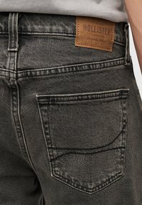 Κοντινό πλάνο προσώπου που φοράει ξεθωριασμένο μαύρο τζιν από τζιν με δερμάτινη ετικέτα Hollister πάνω από την πίσω τσέπη.