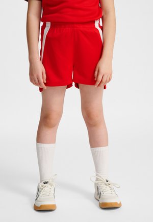 Barn iført røde atletikshorts med hvide sidelister, hvide mellemlange sokker og hvide sneakers, stående mod en ensfarvet baggrund.