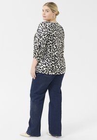 Halenka s leopardím potiskem a rukávy 3/4, s nařasenými manžetami, v kombinaci s modrými širokými džínami a béžovými botami s špičatou špičkou. Bílý pozadí.