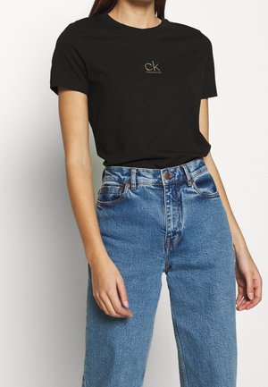 Žena v černém tričku značky Calvin Klein zasunutém do modrých džínů s vysokým pasem, stojící před jednobarevným pozadím.