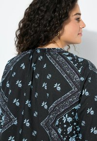 Top de tela negra con un patrón floral en azul claro, con detalles elaborados, pliegues en el escote y un diseño de ajuste relajado.