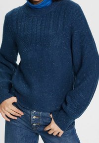 Marineblauer gestrickter Pullover mit strukturiertem Muster und gerippten Bündchen, durchsetzt mit kleinen Farbklecksen. Kombiniert mit hochtaillierten Jeans.