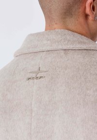 Giacca in pile di colore beige chiaro, con collo a risvolto e logo argentato sul retro. Tessuto morbido con motivi sottili.