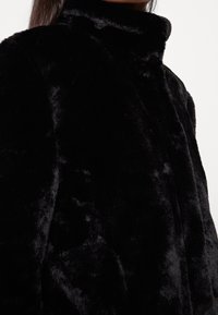 Chaqueta de piel sintética negra con un alto cuello, que presenta una textura suave y un acabado liso, mostrando las costuras y pliegues en el material.