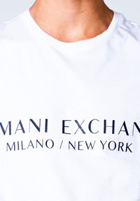 Bílá krátká rukávová tričko s kulatým výstřihem, na kterém je černý text "MANI EXCHANGE MILANO / NEW YORK" v různých sílách písma.