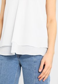 Valkoinen, kerroksellinen pusero, jossa on pehmeä, valuva rakenne. Ylätaso on läpinäkyvä, kun taas alataso on hieman pidempi. Yhdistettyinä sinisiin farkkuihin.