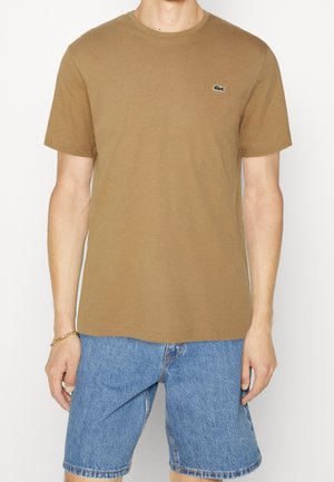 Mężczyzna w prostym brązowym t-shircie z krótkim rękawem, z małym zielonym logo na piersi, oraz w niebieskich dżinsowych szortach.