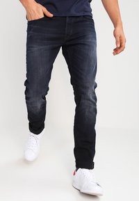 Homme portant un jean slim bleu foncé et des baskets blanches, marchant avec une main dans la poche.