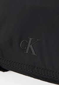 Svart nylonmateriale med en glatt tekstur, med et preget "cK"-logo i nederste hjørne og kontrasterende søm langs kanten.