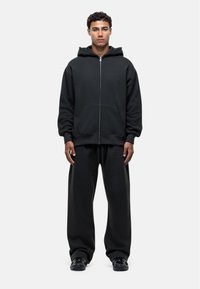 Felpa nera con zip e ampie tasche frontali, accompagnata da pantaloni della tuta neri coordinati. Il tessuto sembra morbido e ha una vestibilità comoda.