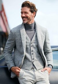 Grau strukturierter Blazer über einem grauen Rollkragenpullover und einer Weste, kombiniert mit hellen Hosen. Auffällige Details sind Knöpfe und ein lässiger Schnitt.