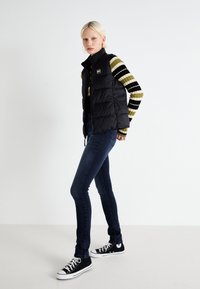 Fekete pufferszövet mellény magas gallérral, csíkos hosszú ujjú póló felett. Slim-fit sötét farmer és fekete sportcipő egészíti ki az öltözetet.