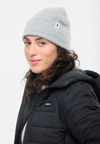 Young woman wearing a grey beanie with a tree logo and a black quilted jacket, looking slightly sideways with a gentle smile.