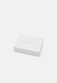 Caja rectangular blanca con el texto dorado "GUESS" en la parte frontal, con una textura suave y un diseño limpio y minimalista.