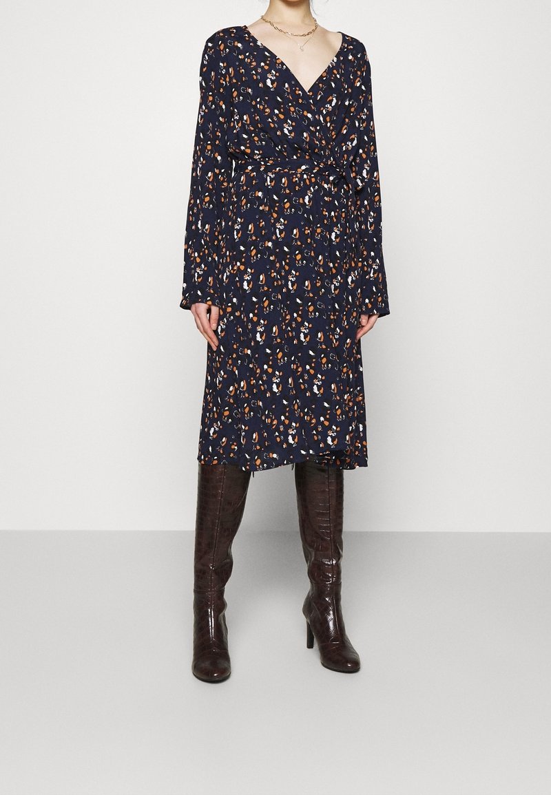 Femme portant une robe portefeuille bleu marine avec des taches orange et blanches, associée à des bottes en cuir marron montant jusqu'aux genoux.