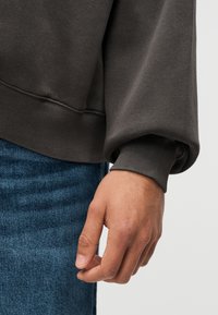 Κοντινό πλάνο του χεριού ενός ατόμου που ξεκουράζεται δίπλα στο ισχίο του, φορώντας σκούρο γκρι φούτερ με ριμπ μανσέτες και μπλε τζιν.