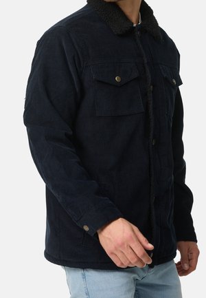 Man draagt een marineblauwe corduroy jas met borstzakken met knopen en een zwarte sherpa-kraag, gecombineerd met lichtblauwe spijkerbroeken.