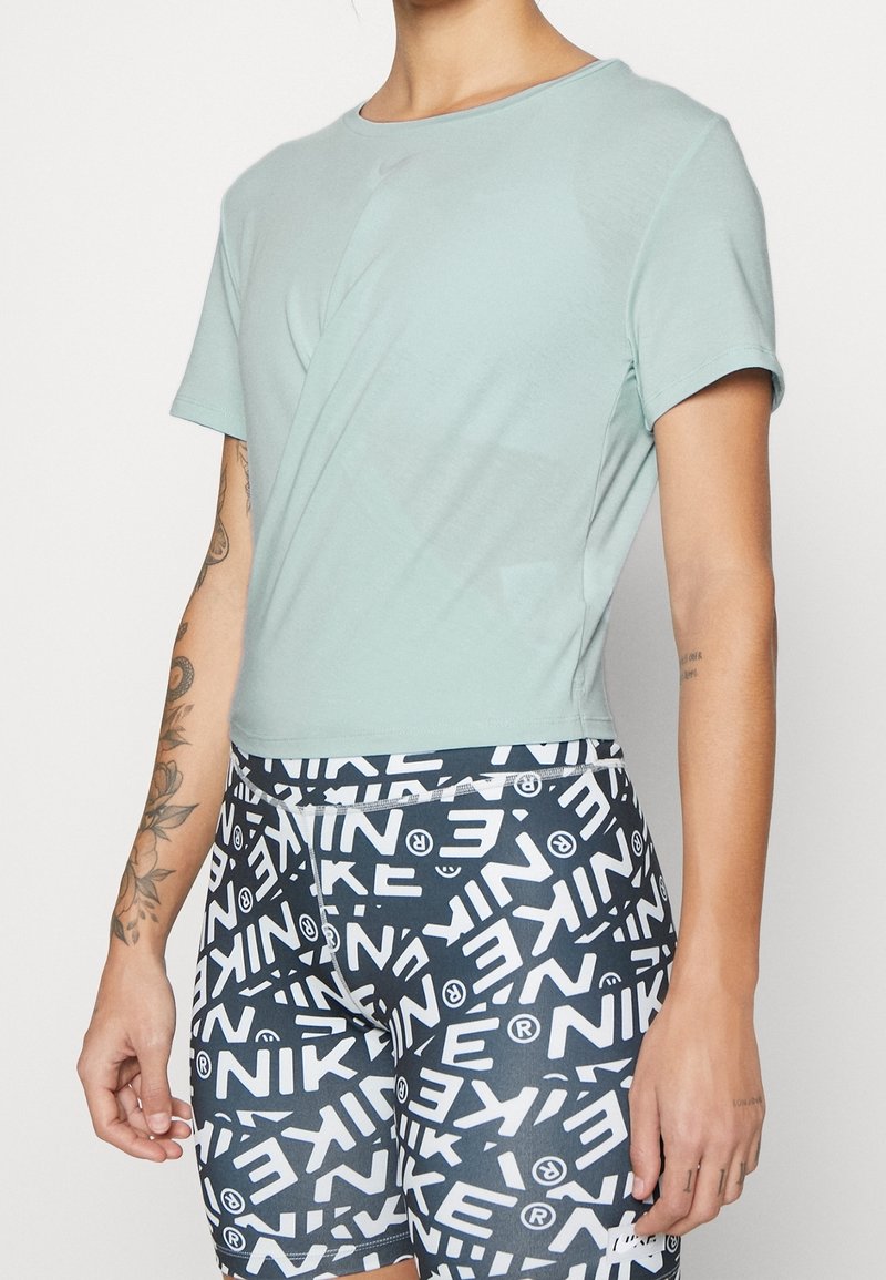 Person som bär en ljusgrön tajt t-shirt och svarta shorts täckta med vita Nike-logotyper, stående mot en enkel vit bakgrund.