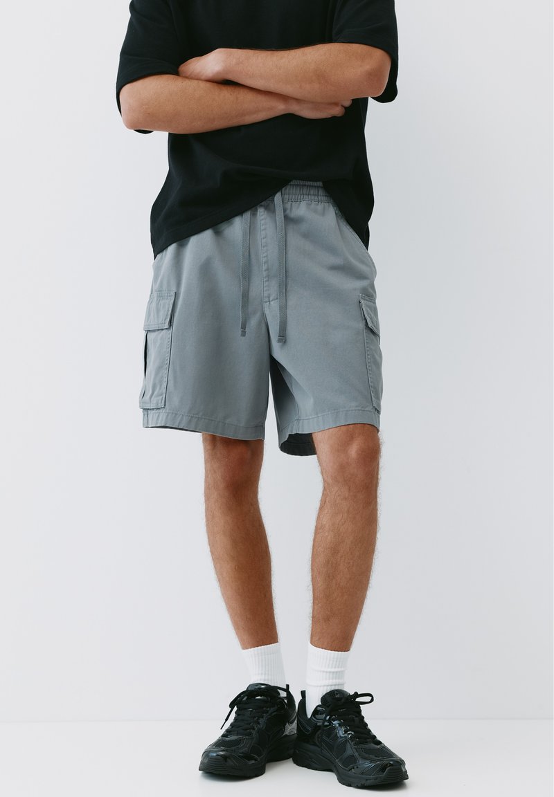 Personne portant un short cargo gris, des baskets noires, des chaussettes blanches et un t-shirt noir à manches courtes, les bras croisés.
