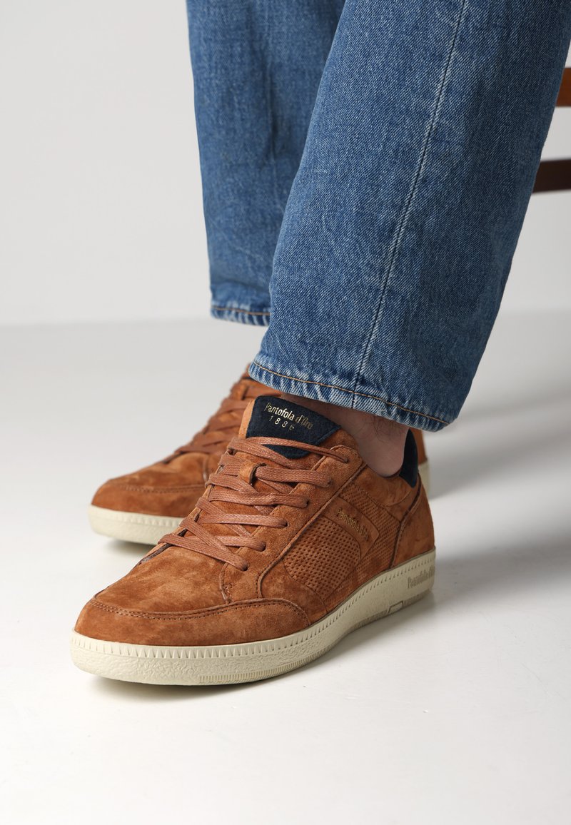 Braune Wildledersneaker mit strukturierten Einsätzen, Schnürdesign und einer cremefarbenen Gummisohle. Teilweise sichtbare blaue Jeans.