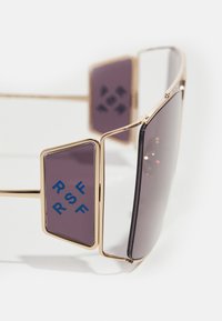 Occhiali da sole con montatura in metallo dorato, lenti scure di grandi dimensioni e dettagli geometrici laterali viola con testo blu "RSF". Design angolare.