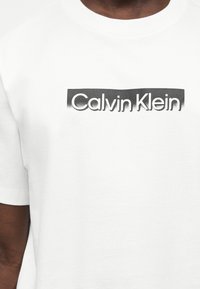 Λευκό T-shirt με το λογότυπο «Calvin Klein» σε μαύρη γραμματοσειρά μέσα σε σκιασμένο ορθογώνιο πλαίσιο στο ύψος του στήθους.
