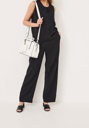Czarny bezrękawnik z marszczonym dekoltem w połączeniu z czarnymi spodniami o szerokich nogawkach. Modelka trzyma białą, usztywnianą torebkę i nosi czarne sandały na słupku.