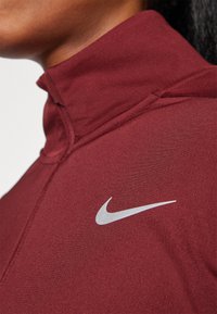 Bordowy sportowy top z półzamkiem, z wysokim kołnierzem i teksturowanym wykończeniem, z białym logo Nike na lewej stronie.