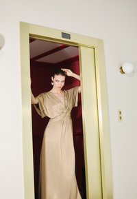 Beige, floor-length dress with a knotted waist and wide sleeves, shown in an elevator with gold accents and a red interior.
