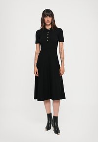 Black ribbed knit dress featuring short sleeves, a collar, and three gold buttons; flared skirt and textured fabric; styled with black heeled boots.