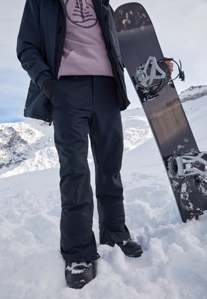 Osoba v lyžařských kalhotách a bundě drží černý snowboard, stojící v zasněženém horském terénu pod zataženou oblohou.
