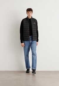 Colete acolchoado preto sobre um suéter preto, combinado com jeans azul claros e sapatos pretos. Textura plana, design minimalista, sem padrões visíveis.